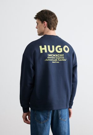 Vīrietis, valkājot tumši zilu sporta džemperi ar dzeltenu "HUGO" tekstu uz muguras, kopā ar zilām džinsu biksēm, stāvot vienkāršas gaiši pelēkas fona priekšā.