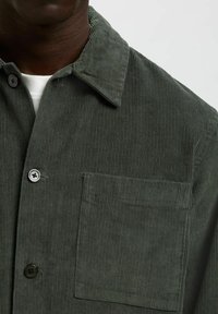 Camicia in corduroy verde scuro con grande tasca sul petto e bottoni di colore diverso, indossata sopra una maglietta bianca a girocollo.