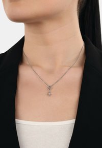 Collana in argento con un piccolo pendente a forma di ancora, indossata sulla pelle nuda con un blazer nero e una maglietta bianca in vista.