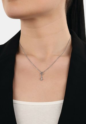Zilveren ketting met een kleine anker hanger, weergegeven op blote huid met een zwarte blazer en een witte top zichtbaar.