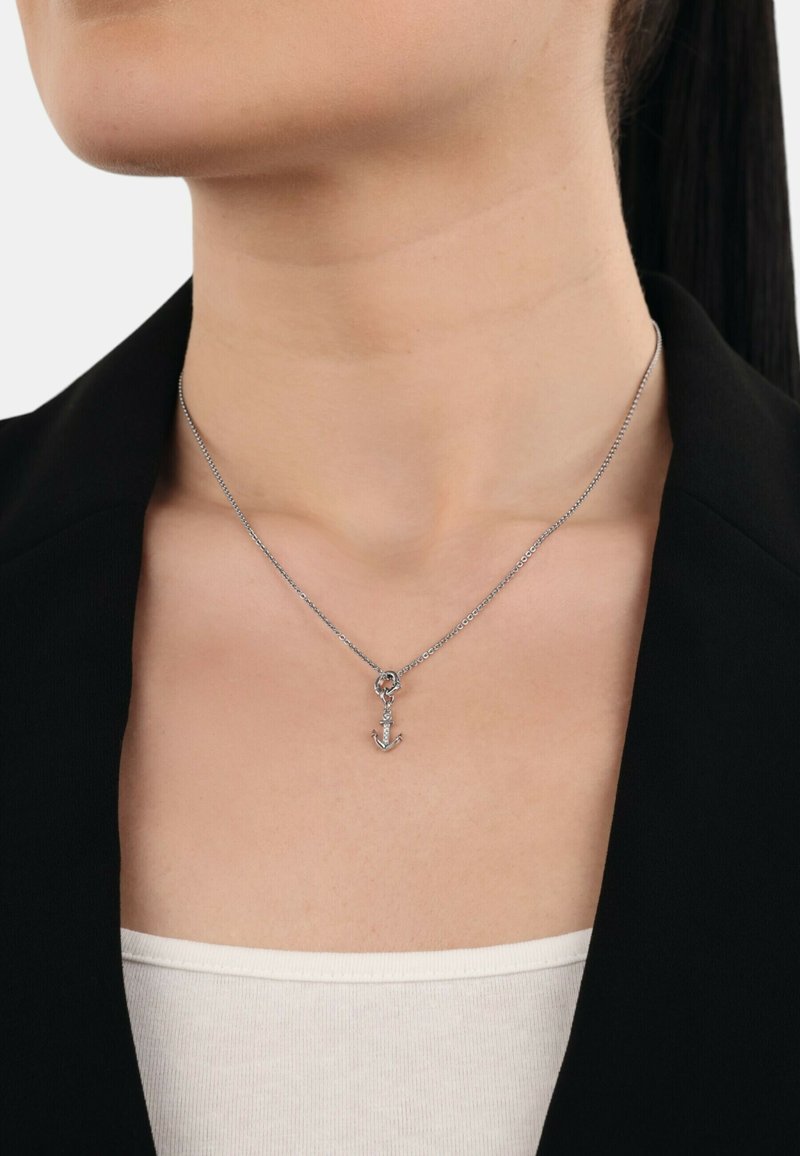 Collana in argento con un piccolo pendente a forma di ancora, indossata sulla pelle nuda con un blazer nero e una maglietta bianca in vista.