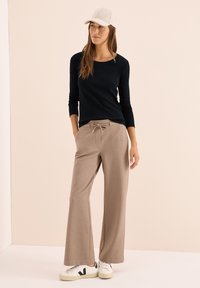 Zwart lang mouwtop, beige wijde broek met trekkoord en witte sneakers met zwarte accenten. Model draagt een lichtgekleurde pet.