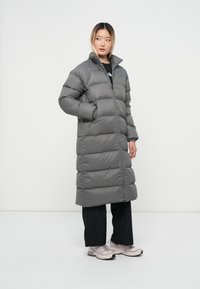 Grijze, gewatteerde lange jas met een hoge kraag, voorzien van verticale quilted secties en een voorkant met rits. Gekozen boven zwarte broek en lichte sneakers.