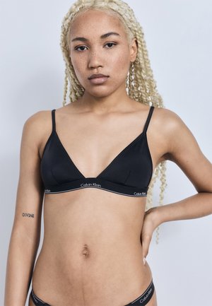 Szőke, fonott hajú nő fekete Calvin Klein melltartóban és fehérneműben, egy kézzel a csípőjén áll, egyszerű háttér előtt.