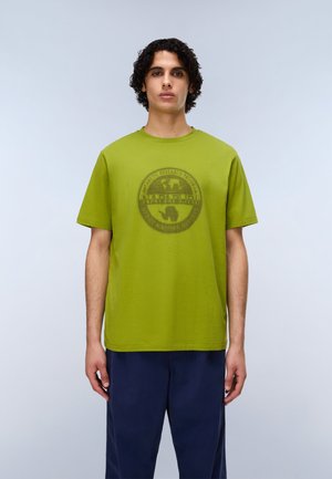 Zelené bavlnené tričko s okrúhlym výstrihom a krátkymi rukávmi, na prednej strane s tmavým grafickým logom. Nosí sa s tmavomodrými nohavicami.