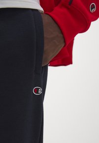 Los pantalones de chándal de lana en color azul marino presentan un bolsillo lateral y un logotipo bordado con detalles en rojo y blanco. La imagen en primer plano muestra una mano descansando en el bolsillo.