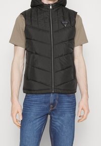 Schwarze Steppweste mit Kapuze, ausgestattet mit einem Frontreißverschluss und Zickzacknähten. Trägt man über einem beigen T-Shirt und kombiniert mit blauen Jeans.