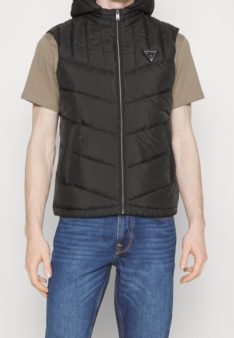 Schwarze Steppweste mit Kapuze, ausgestattet mit einem Frontreißverschluss und Zickzacknähten. Trägt man über einem beigen T-Shirt und kombiniert mit blauen Jeans.