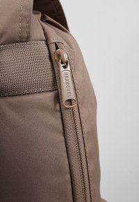 Gros plan sur un sac à dos en tissu beige dotée d'une fermeture éclair verticale avec un curseur métallique gravé du nom de la marque Herschel.