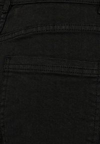 Crni teksas blizu, s glatkom teksturom i detaljem ušivenih džepova. Materijal prikazuje suptilan dijagonalni keper uzorak.