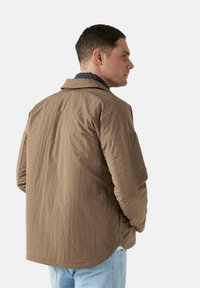 Giacca marrone con texture e colletto standard, caratterizzata da una vestibilità rilassata e tasche laterali, indossata sopra una camicia blu navy.