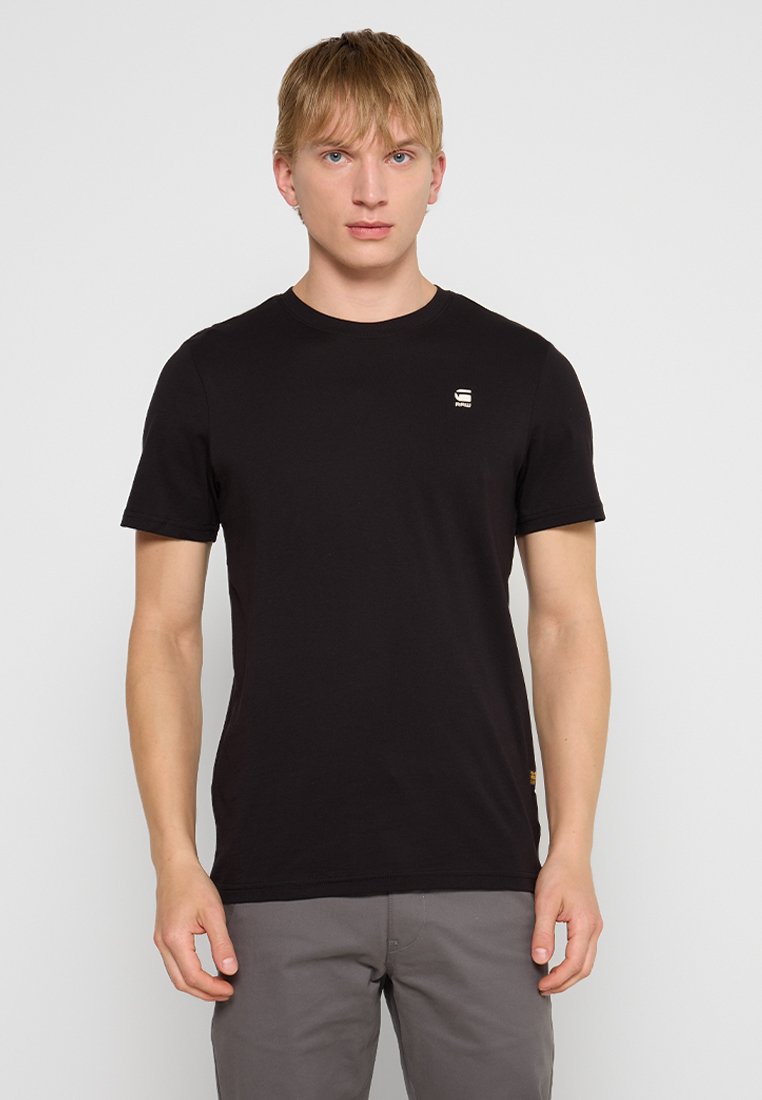 Młody mężczyzna z blond włosami, ubrany w zwykły czarny t-shirt z krótkim rękawem i szare spodnie, stoi na tle jasnoszarym.
