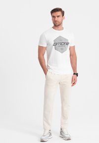 Weißes Baumwoll-T-Shirt mit grauem hexagonalem Logo, auf dem "ombre casual style 1985" steht, kombiniert mit hellbeigen Hosen und grauen Sneakers.