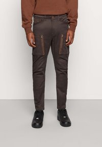 Cargo kalhoty v tmavě hnědé látce s kapsami na zip, slim fit, texturovaný povrch, zesílené detaily na kolenou, zkombinované s černými teniskami.