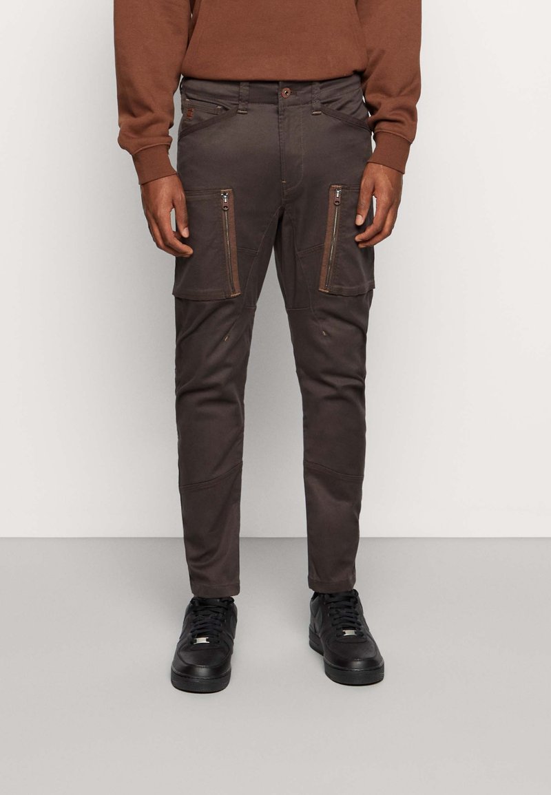 Cargo kalhoty v tmavě hnědé látce s kapsami na zip, slim fit, texturovaný povrch, zesílené detaily na kolenou, zkombinované s černými teniskami.