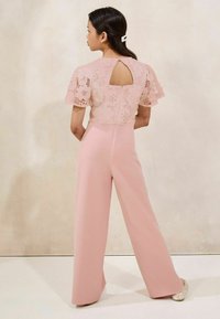 Femme avec des cheveux foncés coiffés d'une pince à cheveux en perles portant une combinaison en dentelle rose avec des pantalons à jambes larges, se tenant devant un mur beige, de dos.