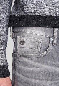 Jeans en denim gris avec un pull à texture par-dessus. Comprend une poche pour les pièces de monnaie et des garnitures en métal, soulignant un style décontracté.