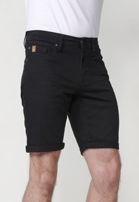 Mies, joka pukeutuu mustiin käännettyhelmaisiin denim-shortsseihin ja valkoiseen sisätakkiin, näkyy vyötäröstä polviin yksinkertaista taustaa vasten.