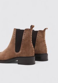 Botines de ante marrón con paneles elásticos negros, perforaciones decorativas y pequeños remaches dorados en las costuras, que cuentan con un tacón bajo negro.