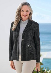 Schwarzer Blazer aus einem strukturierten Stoffmuster mit einem einzelnen Knopfverschluss, zwei Reißverschlusstaschen und einer schlanken Passform, kombiniert mit einem hellgrauen Rollkragenpulli.