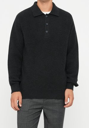 Homme portant un pull en maille noir texturé manches longues avec une patte à trois boutons et un pantalon à carreaux gris.