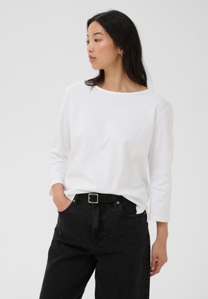 Femme aux longs cheveux noirs porte une chemise blanche à manches longues et un pantalon noir avec ceinture, debout, une main dans la poche, regardant sur le côté.