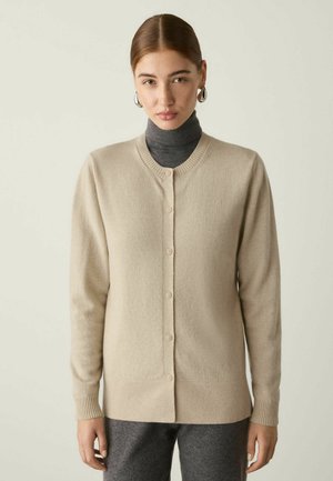 Kvinde med brunt hår iført sølvfarvede øreringe i ringe, beige cardigan med knapper over mørkegrå rullekrave og mørkegrå bukser.