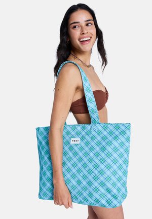 Jonge vrouw in een bruin bikinibovenstuk, glimlachend, met een grote blauwgroen geruite draagtas met het label "Roxy" over haar schouder.