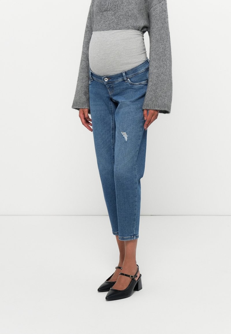 Personne enceinte portant un jean de maternité bleu avec une bande élastique grise sur le ventre, un pull gris et des chaussures à talons noires, debout sur un sol blanc.