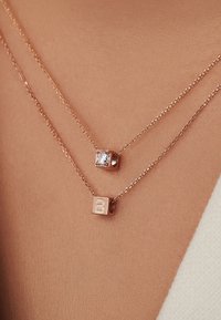 Des colliers en or rose superposés ; l'un présente une pierre précieuse carrée transparente, l'autre a un charme en forme de cube avec la lettre "B".