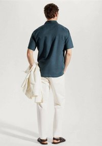 Lyhythihainen, tummanviherhtävä paita yhdistettynä valkoisiin housuihin. Malli pitää kädessään vaalean väristä takkia ja jalassa on tummat sandaalit. Minimalistinen muotoilu.