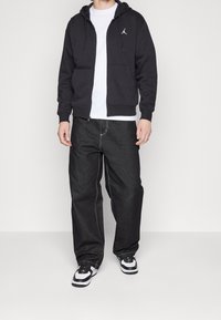 Uomo che indossa una felpa nera con zip e logo Jordan, una maglietta bianca, jeans neri larghi e sneaker bianco e nere, in piedi su uno sfondo bianco.