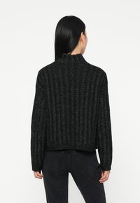 Vrouw met lang zwart haar, gekleed in een zwarte ribgebreide trui en donkere jeans, gezien vanaf de rug tegen een effen achtergrond.