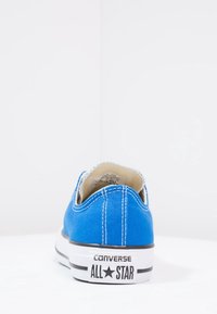 Blå canvas sneaker med vit sömnad, låg design, svart gummisulor och svart "CONVERSE ALL STAR"-branding på hälen.