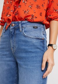 Jeans de denim azul claro con cintura alta, que cuentan con un bolsillo frontal, cierre de botón y una pequeña etiqueta negra con el logo en la cintura.