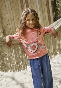 Roze sweatshirt met hartontwerp en versieringen, gecombineerd met losse blauwe broek met zakken. Kind staat tegen een fringed achtergrond.