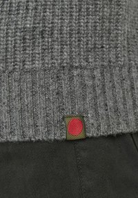 Detail sivého pleteného okraja svetra s malou zelenou visačkou s červeným kruhom, nosený na tmavých látkových nohaviciach.