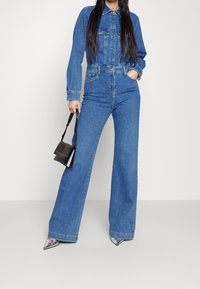 Jeansjumpers med långa ärmar, knäppning fram, två bröstfickor och utsvängda ben i mellanblått. Modell som håller en liten svart väska.