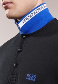 Black polo shirt with a blue and white knit collar. Features black buttons and a blue embroidered "BOSS" logo on the chest.