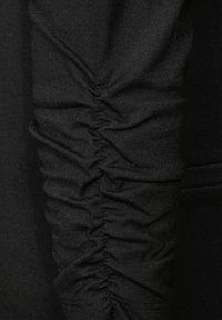 Närbild av svart tygärm med rynkad, samlad sömnad som skapar texturerade veck längs hela längden.