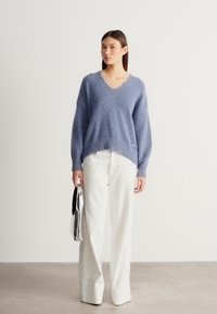 Ανοιχτό γαλάζιο πλεκτό πουλόβερ με V-λαιμόκοψη, μακριά μανίκια και ριμπ τελείωμα, συνδυασμένο με λευκό παντελόνι φαρδιά μπατζάκια και ασημί τσαντάκι φάκελο.