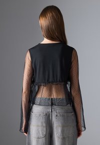 Fekete cropped top áttetsző, pöttyös hálós betéttel és harangujjakkal. A hátsó részén fodros szegély található. Szürke, magas derekú farmerral párosítva.