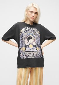 Νεαρό άτομο με μακριά ξανθά μαλλιά φοράει μαύρο φαρδύ μπλουζάκι με σχέδιο γιν και γιανγκ και ήλιο, συνδυασμένο με κίτρινες ριγέ παντελόνες.