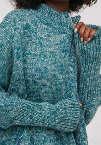 Sweter zapinany na zamek w kolorze szmaragdowym i szarym, w melanzowany wzór, z wysokim kołnierzem, długimi rękawami i prążkowanymi mankietami, noszony przez osobę z ciemnymi kręconymi włosami.