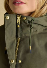 Jasnozielona kurtka z zamek błyskawicznym z przodu, złotymi elementami oraz regulowanym kapturem. Posiada zapięcia na guziki i teksturowany materiał.
