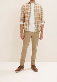 Brązowa koszula w kratę na jasnokrémowym swetrze, w zestawieniu z jasnobeżowymi spodniami i brązowymi sznurowanymi butami na gumowej podeszwie. Prosty i warstwowy design.