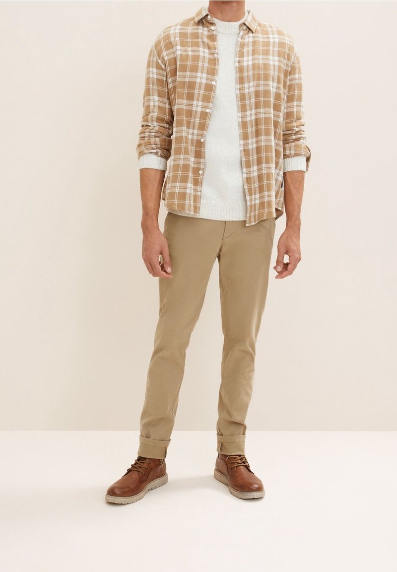 Brązowa koszula w kratę na jasnokrémowym swetrze, w zestawieniu z jasnobeżowymi spodniami i brązowymi sznurowanymi butami na gumowej podeszwie. Prosty i warstwowy design.