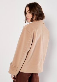 Donna con capelli corti castani che indossa un cappotto di lana beige e pantaloni marroni, mentre guarda oltre la spalla su uno sfondo bianco semplice.