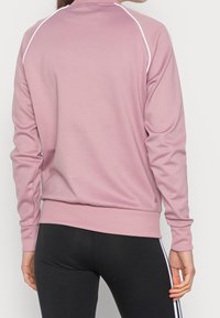 Kvinna som bär en dammig rosa långärmad träningsjacka med vit piping och svarta leggings med vita sidostreck, stående med ryggen mot kameran.