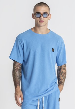 Mężczyzna z tatuażami nosi niebieską teksturowaną koszulkę i pasujące spodenki, do tego duże okulary przeciwsłoneczne z niebieskimi szkłami oraz mały kolczyk.